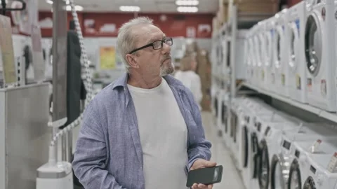 An elderly man walks through a hypermarket and looks at washing machines Stock Footage 251735296