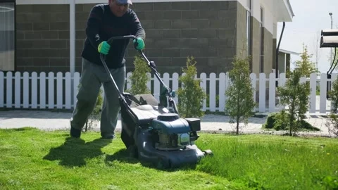 Elderly man in work gloves and clothes pushes lawnmower to cut an overgrown lawn Stock Footage 275076050