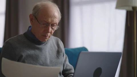 Elderly man is working in home, viewing papers and using laptop for typing Stock Footage 153530225