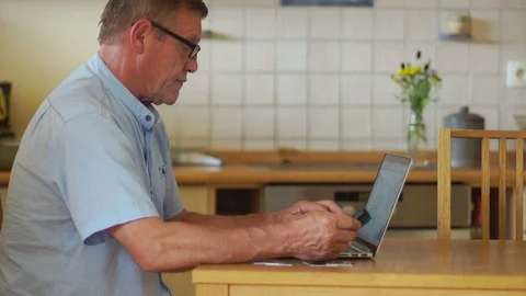 An elderly man works with a computer and phone, sitting at a table in his Stock Footage 91976321