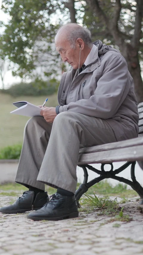 Elderly man writing on a notepad while sitting on a bench Stock Footage 327230197