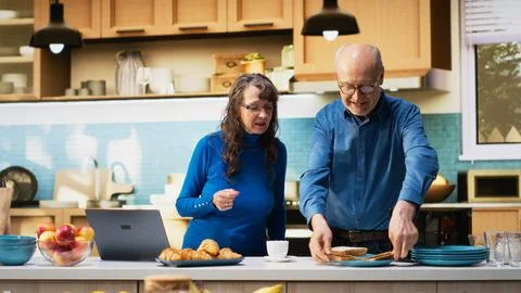 Elderly married pair navigating websites on laptop in home kitchen Fotos Stock