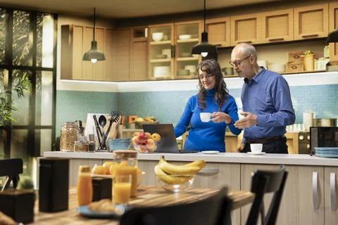 Elderly married pair navigating websites on laptop in home kitchen Fotos Stock