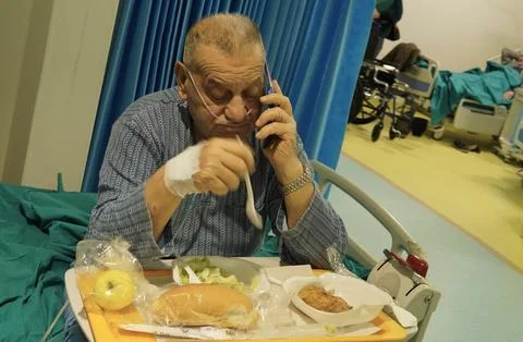 An elderly patient eats and uses a smartphone in a hospital room. 스톡 사진