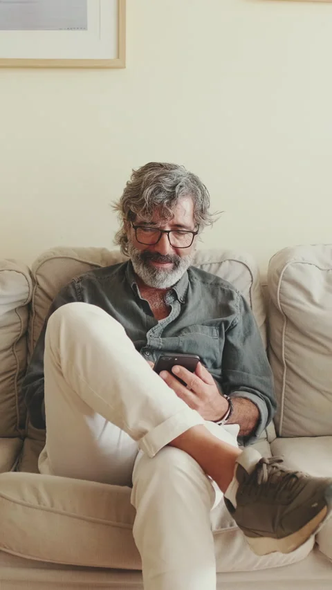 Elderly positive man using mobile phone while sitting on sofa at home Stock Footage 229507970