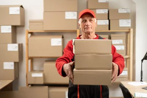 Elderly post office worker standing with stack of parcels near storage rack.. Stock Photos