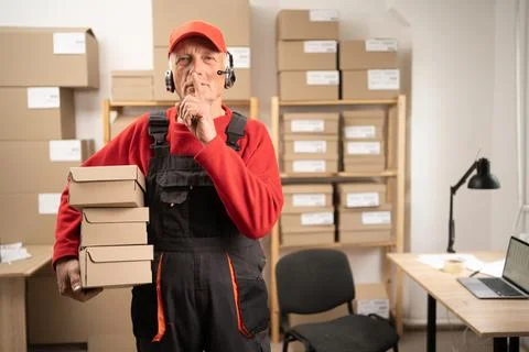 Elderly postal worker with wireless headset showing silence gesture and looki Stock Photos
