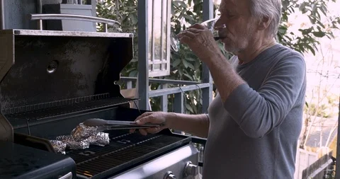 Elderly senior man drinking red wine turning potatoes on a bbq Stock Footage 86004115