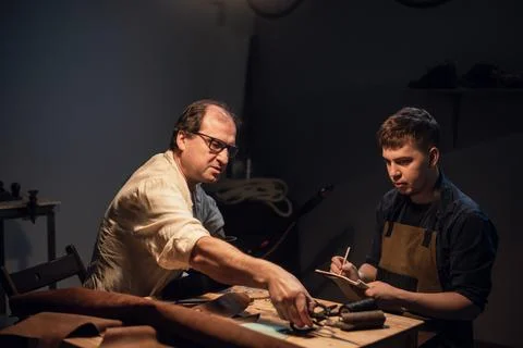 An elderly shoemaker and his apprentice create shoes by hand in their workshop Stock Photos
