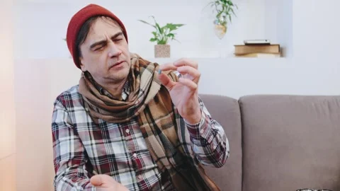 An elderly sick man in a red cap and plaid scarf focuses on medicine in a jar in Stock Footage 301572390