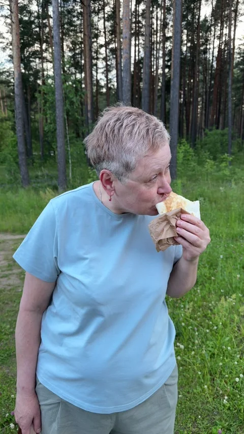 An elderly woman eats shawarma with beef meat outdoors in the forest 库存影片 338179315