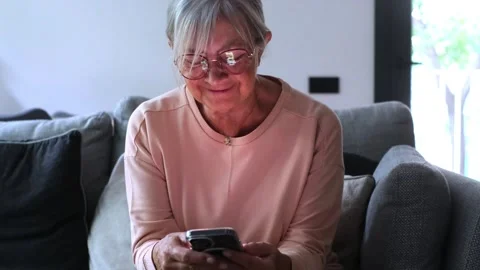 Elderly woman with glasses smiling while using her smartphone, sitting Stock Footage 344782813