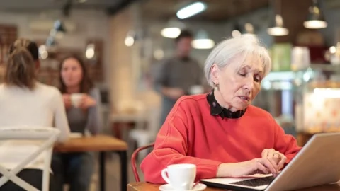 Elderly woman sitting at table, using laptop and enjoing cup of tea in coffee Stock Footage 343715455
