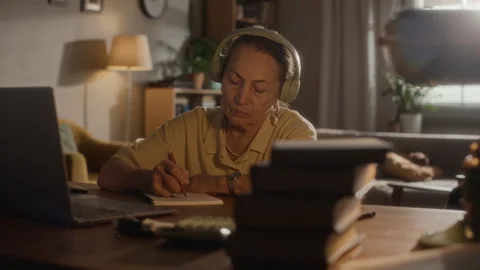 Elderly Woman Taking Notes while Using Laptop with Headphones Indoors Stock Footage 335756084