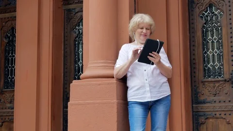 Elderly woman using tablet computer with earphones. Vintage building in the back Stock Footage 78880703