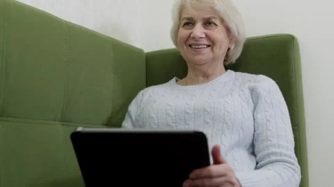 Elderly woman using tablet device on green sofa with focused concentration Stock Footage 322812753