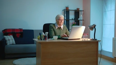 An elderly woman works at a computer while sitting at a table in the evening Stock Footage 128758432