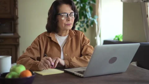 Elderly woman write note on notebook and check information with laptop. Female Stock Footage 156755741