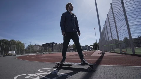 Electric Longboard man in blue jacket and hat ride on red sport stadium with Stock Footage 90815740