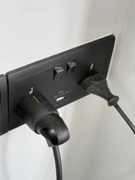 Electrical outlet with two plugged-in cords on a white wall, close-up shot Stock Photos