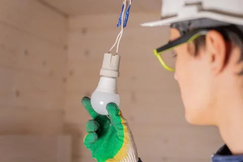 Electrician changes energy-saving light bulb. Worker in a white helmet checki Stock Photos
