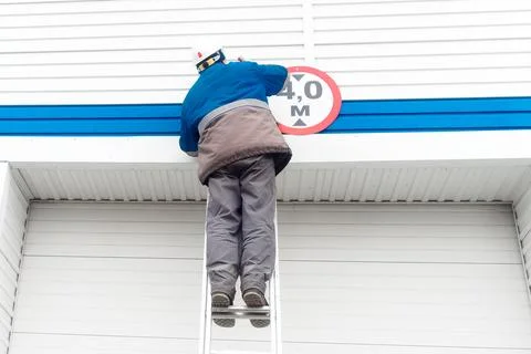 An electrician in a hard hat changes a light bulb in a ceiling lamp near an Stock Photos
