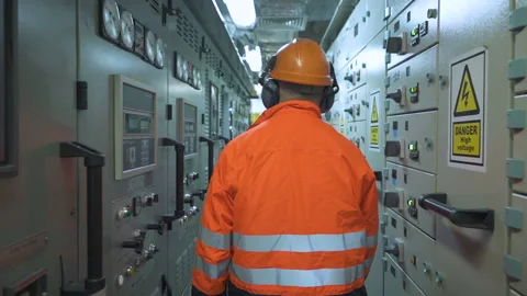 Electrician walks down corridor between electrical panels. Man in orange jacket Stock Footage 196383308