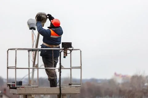 An electrician in work clothes changes street lamps while standing on a high  Stock Photos