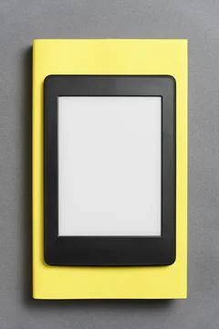 Electronic reader with empty screen over a closed yellow book, gray background Stock Photos