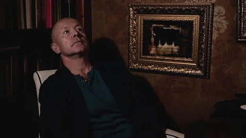 An elederly man is thinking something over in his study with paintings. Stock Footage 120486886