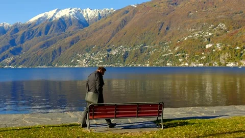 Elegant Man Sitting on a Bench on Alpine Lake Maggiore with Mountain View Видео 164895543