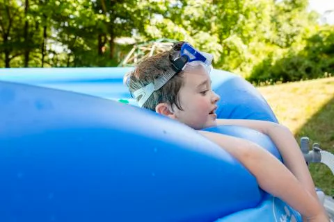 Elementary Age Boy Wearing Goggles on Forehead Relaxes on Side of Pool 스톡 사진