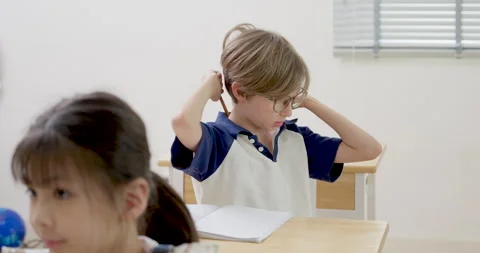 Elementary boy with glasses thinks, adjusts hair, raises pencil during classroom Stock Footage 341929411
