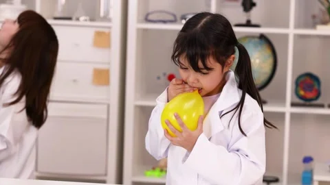 Elementary School Girl in Lab Coat Blowing Up Yellow Balloon Stock Footage 344942658