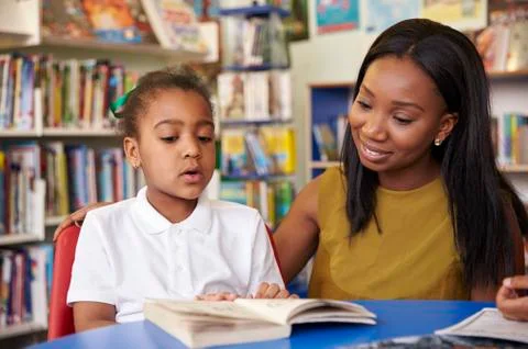 Elementary School Pupil Reading In Library With Teacher Foto stock