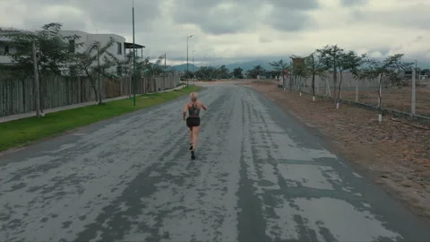 Elevated back view of female runner approaching distant buildings on paved road Stock Footage 339623667