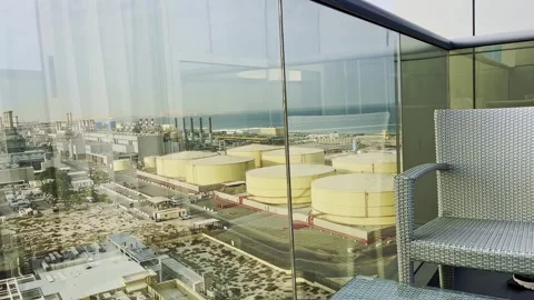 Elevated balcony view of coastal refinery tanks beside woven chair and glass Stock Footage 345437497