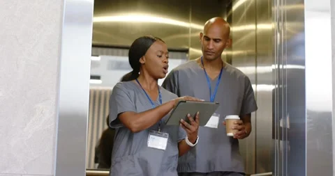 Elevator opening, two staff in scrubs, woman tapping tablet and man holding 库存影片 339651633