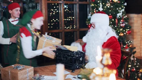 Elves bring gift boxes to Santa Claus, who sits at a table Stock Footage 118859910