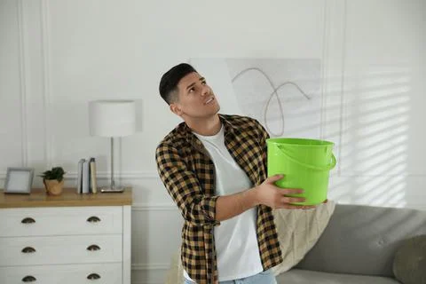 Emotional man collecting water leaking from ceiling in living room. Damaged r Stock Photos