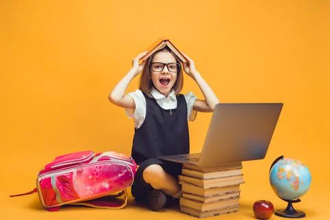 Emotional pupil sits behind a stack of books and laptop holds book on head. Kids Stock Photos