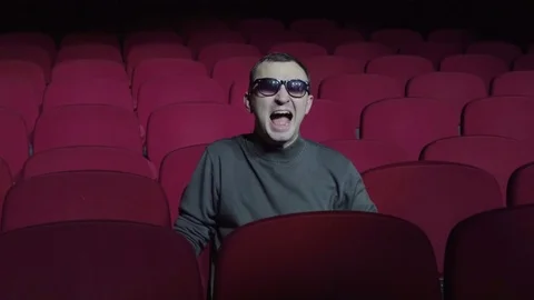 Emotional single man is screaming at comfortable red chairs in dark cinema Stock Footage 83950317