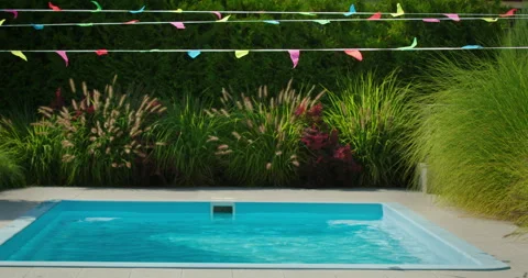 Empty backyard pool with colorful party flags above, surrounded by tall green Stock Footage 308940980