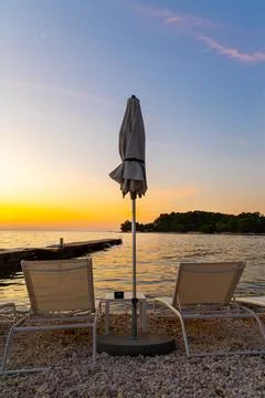 Empty beach chairs facing sunset by sea. Two lounge chairs and a closed umbrella Stock Photos