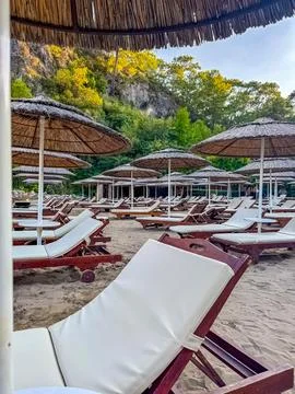 Empty beach sun-bed under straw umbrellas, surrounded by green and rocky cliffs. Stock Photos