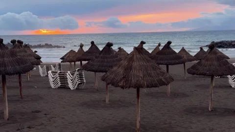 Empty beach with thatched straw parasols and stacked sun loungers at sunset by t Stock Footage 328847801