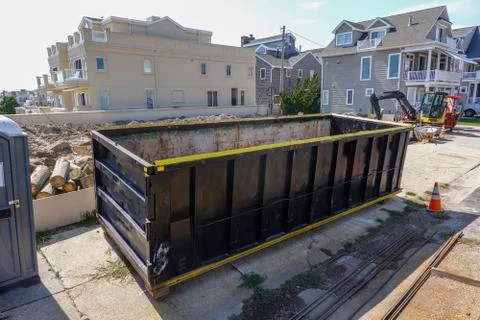 An empty black dumpster is seen at a construction site near a backhoe Stock Photos