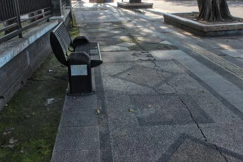 An empty black metal bench on a cracked concrete sidewalk in an urban area. Stock Photos