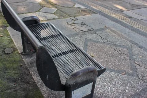An empty black metal bench on a cracked concrete sidewalk in an urban area. Stock Photos