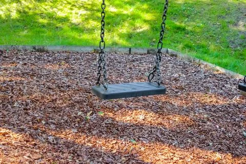 An empty black swing hangs on long steel chains over a wood-chip surface in.. Stock Photos